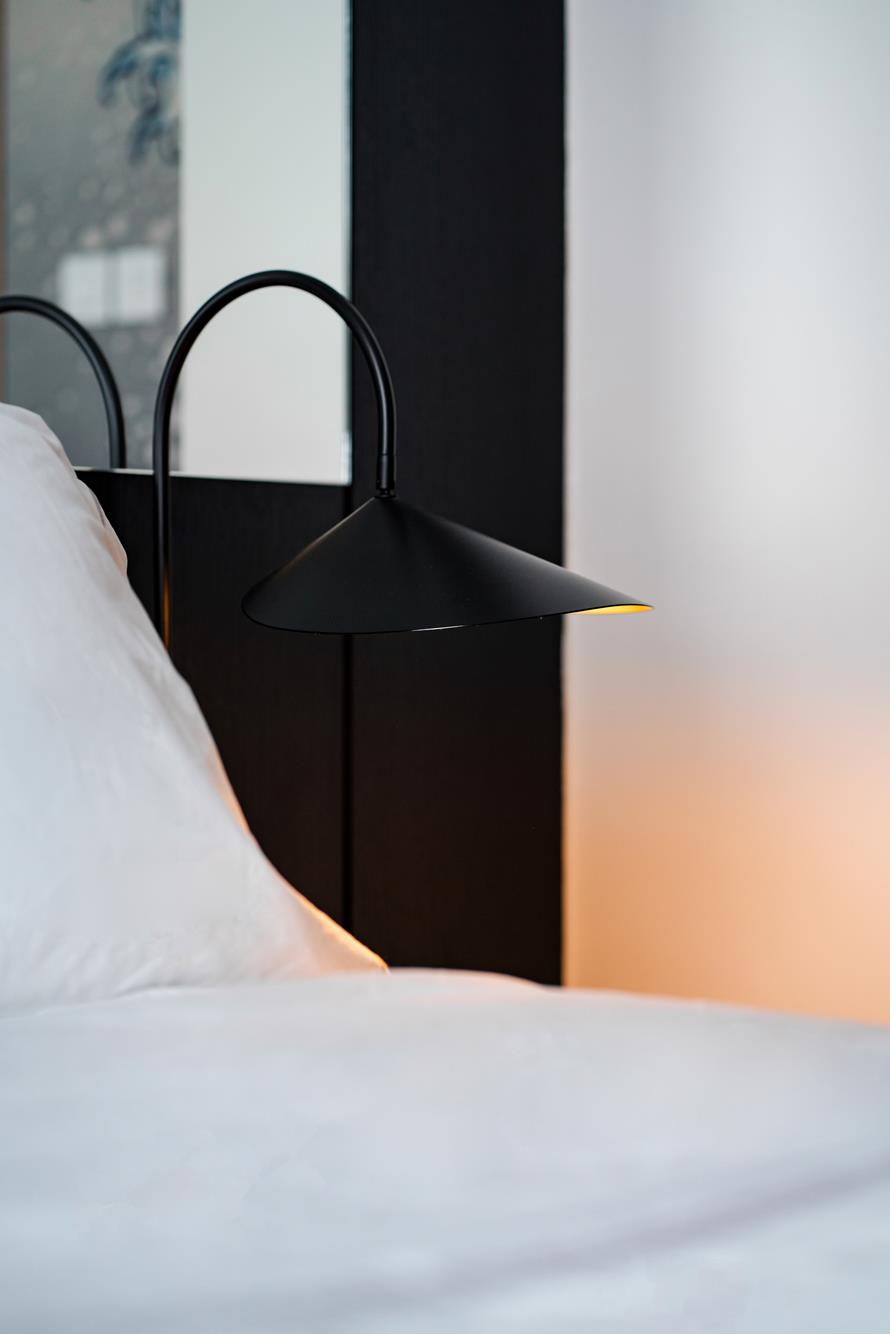 A black wall lamp arcing over white linen against a dark headboard