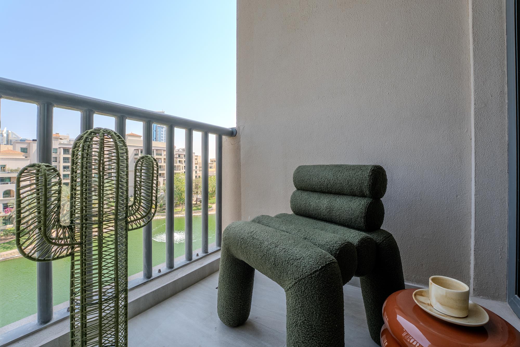 A green bouclé chair on the balcony beside a woven cactus, the lake and low-rise blocks beyond